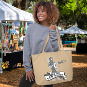 A woman with curly hair is standing in front of a tent, holding a beige tote bag with a graphic design on it.