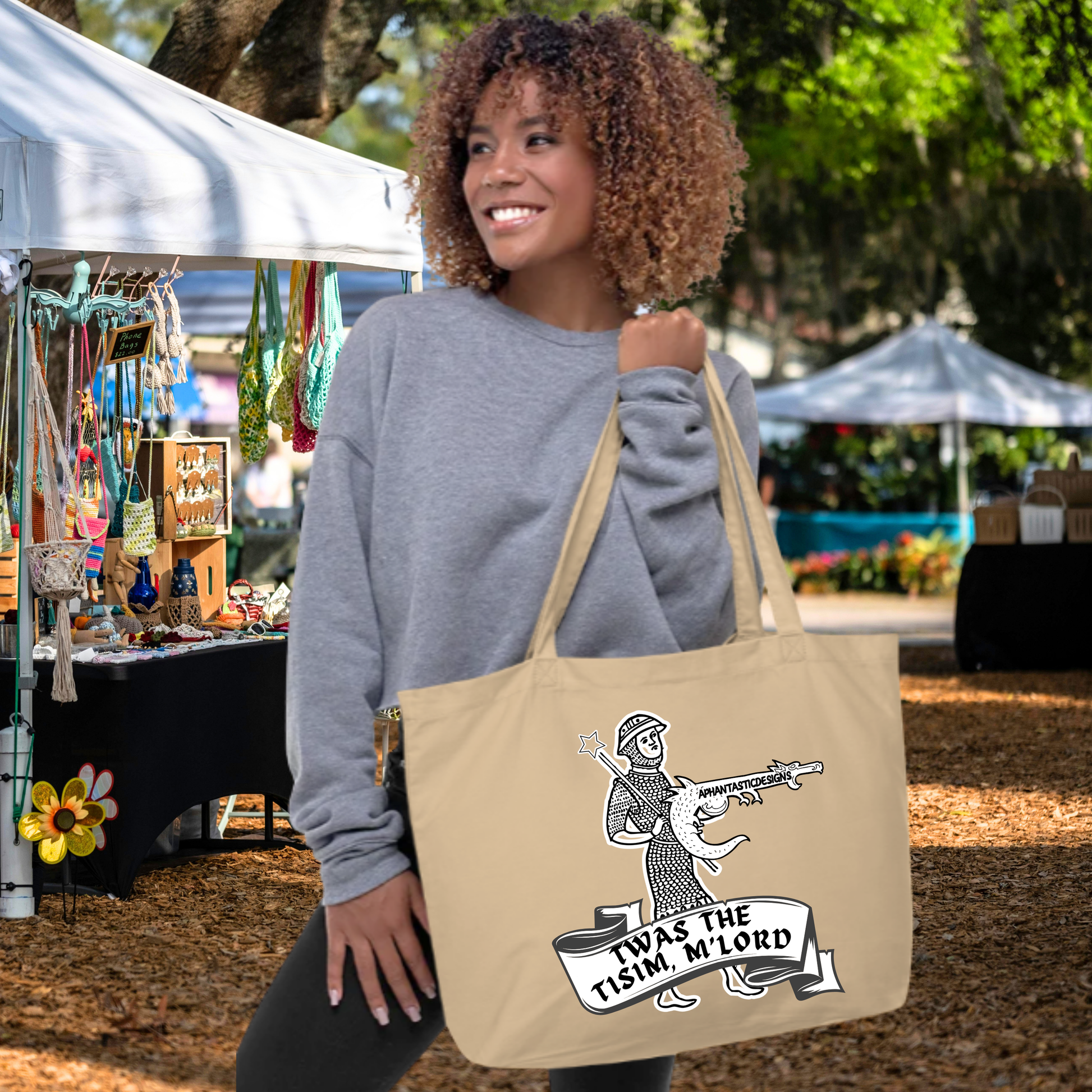 A woman with curly hair is standing in front of a tent, holding a beige tote bag with a graphic design on it.