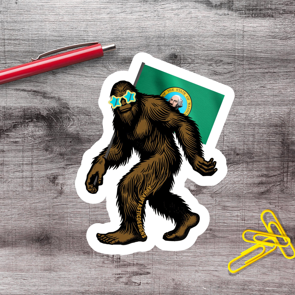 a sticker of a cartoon Sasquatch wearing sunglasses and holding a flag, along with a red pen and yellow paper clips on a wooden surface.
