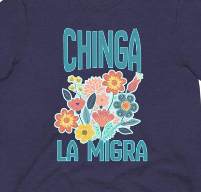 A dark blue t-shirt with a graphic design featuring colorful flowers and the text "CHINGA LA MIGRA" (which means "Migration" in English).