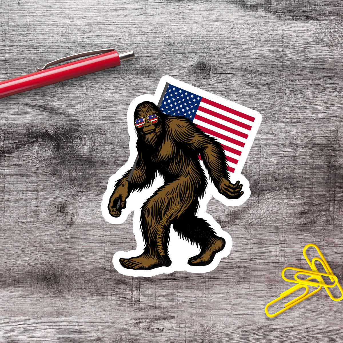 a sticker of a Sasquatch wearing sunglasses and carrying an American flag, along with a red pen and yellow paper clips on a wooden surface.