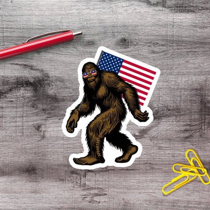 a sticker of a Sasquatch wearing sunglasses and carrying an American flag, along with a red pen and yellow paper clips on a wooden surface.