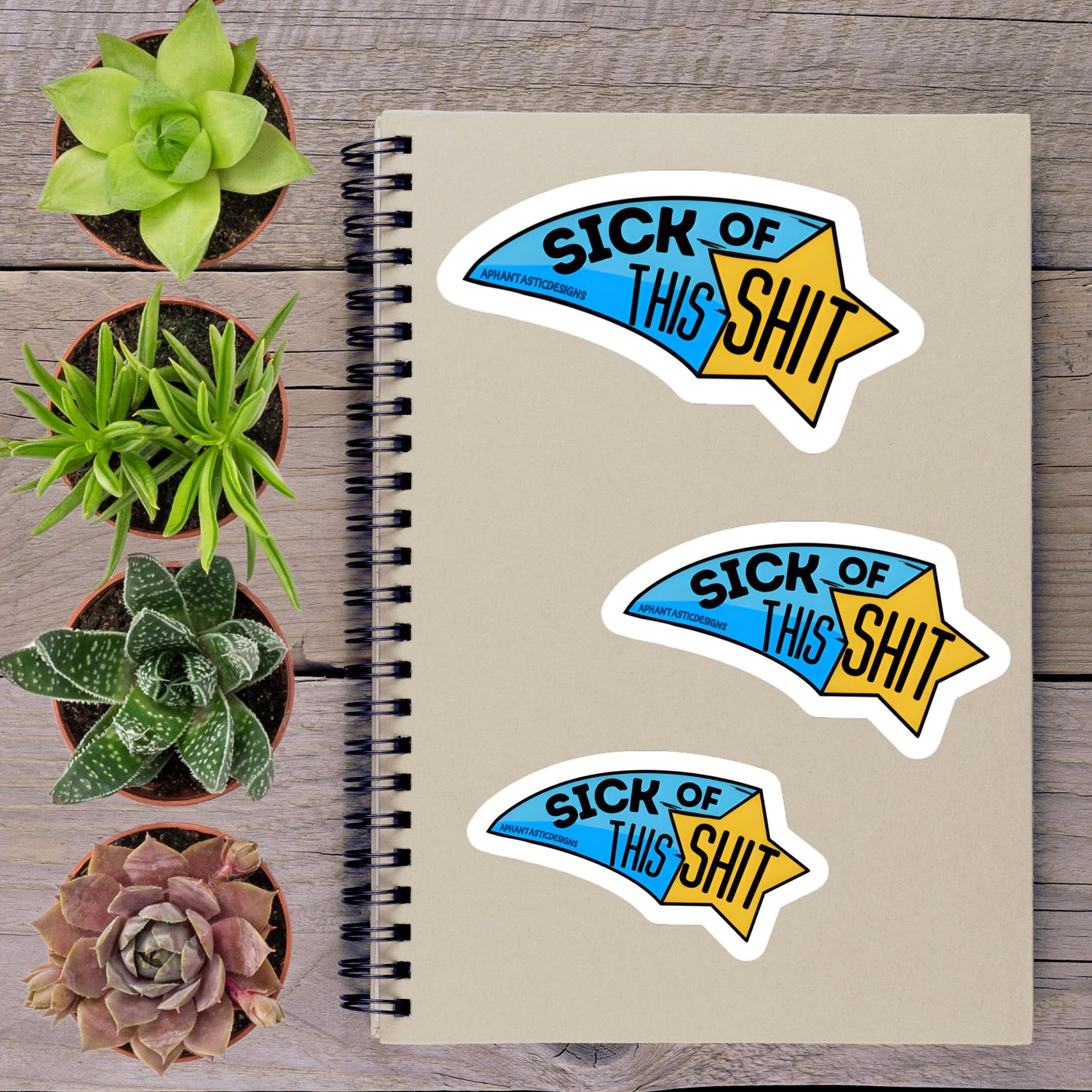 a spiral-bound notebook with three stickers on it, along with several potted plants arranged on a wooden surface.