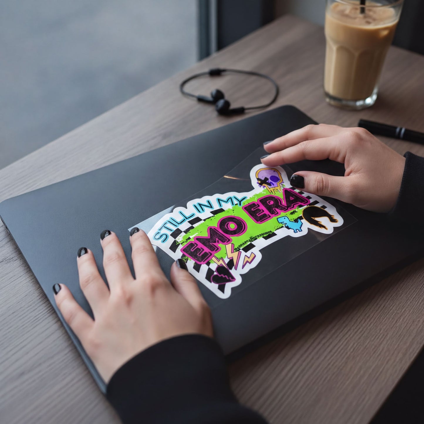 A person's hands are holding a laptop with a sticker on it, which says "STILL IN MY EMO ERA".