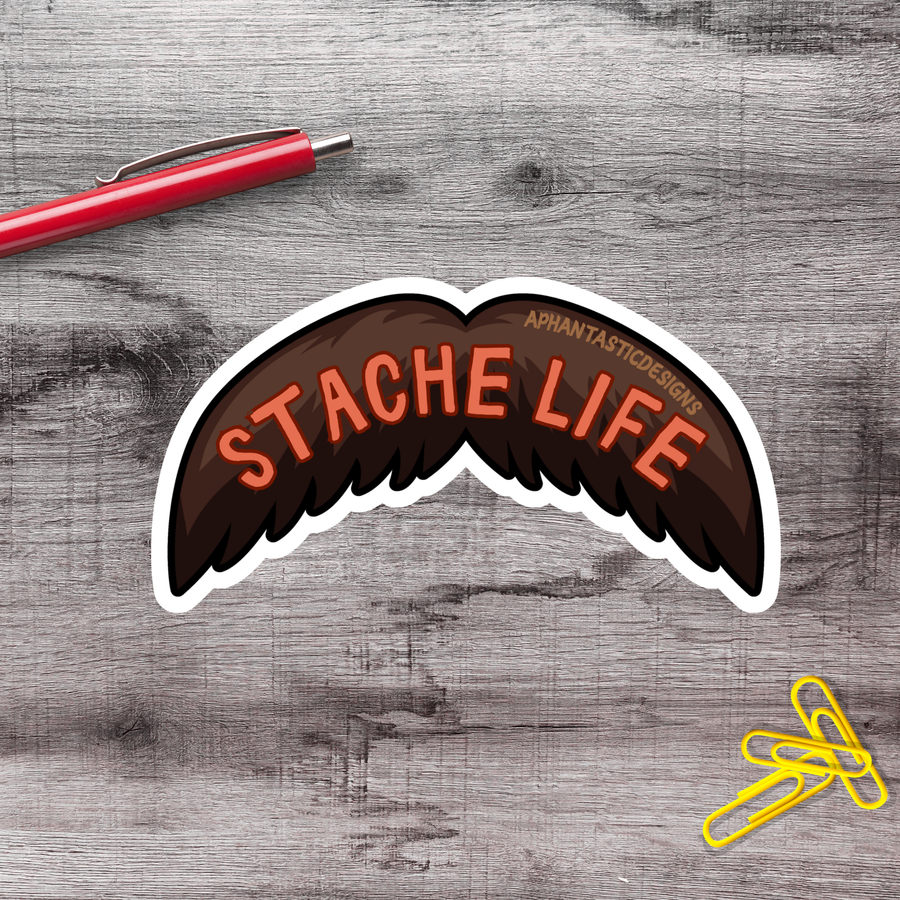 a sticker with the text "STACHELIFE" and a stylized mustache, along with a red pen and a pile of yellow paper clips on a wooden surface.