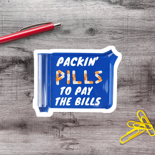 a blue sticker with the text "PACKIN' PILLS TO PAY THE BILLS" printed on it, placed on a wooden surface alongside a red pen and yellow paper clips.