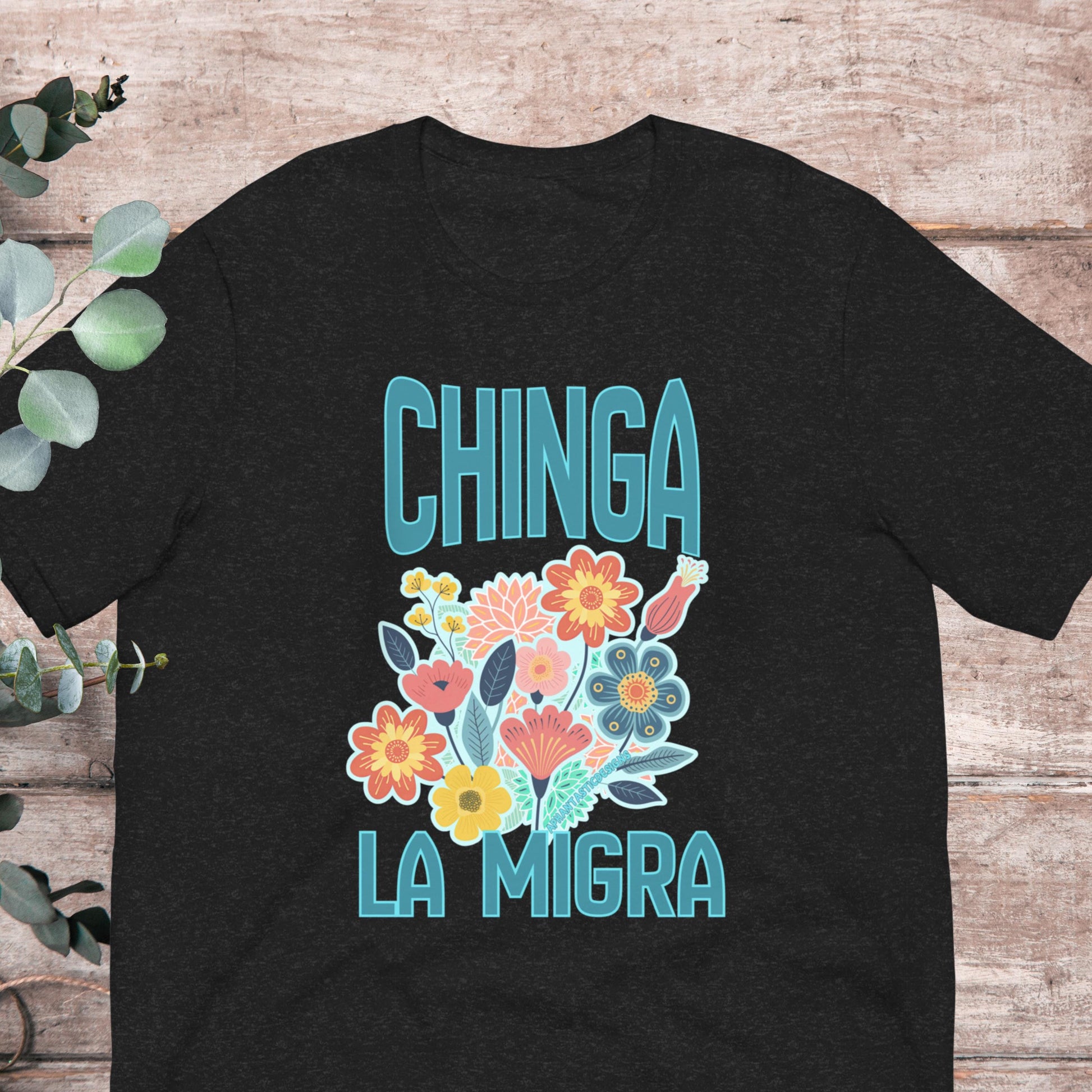 A black t-shirt with a colorful graphic design featuring flowers and the text "CHINGA LA MIGRA" (Spanish for "Migration") printed on it.