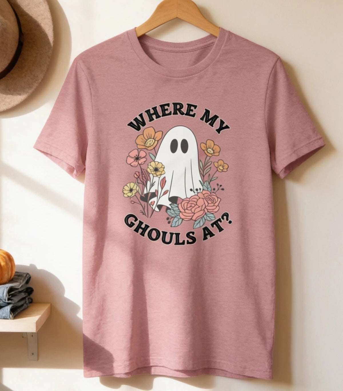 a pink t-shirt hanging on a wooden hanger, with a hat and two pumpkins placed on a shelf below it.