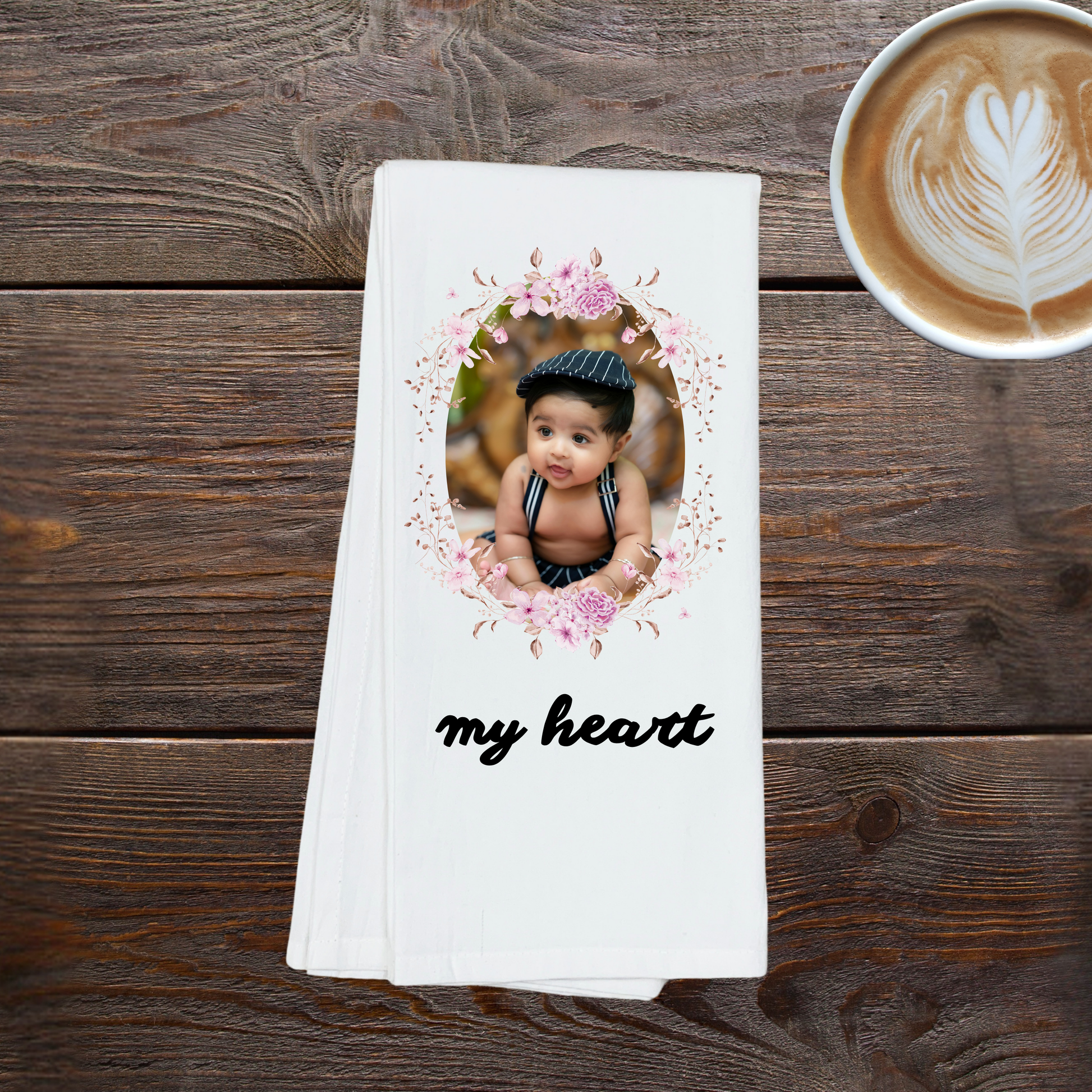 A white towel with a photo of a baby and the text "my heart" printed on it, placed on a wooden surface next to a cup of coffee.