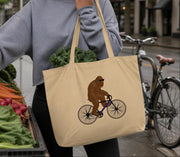 A person is holding a beige tote bag with a brown cartoon gorilla riding a bicycle printed on it.