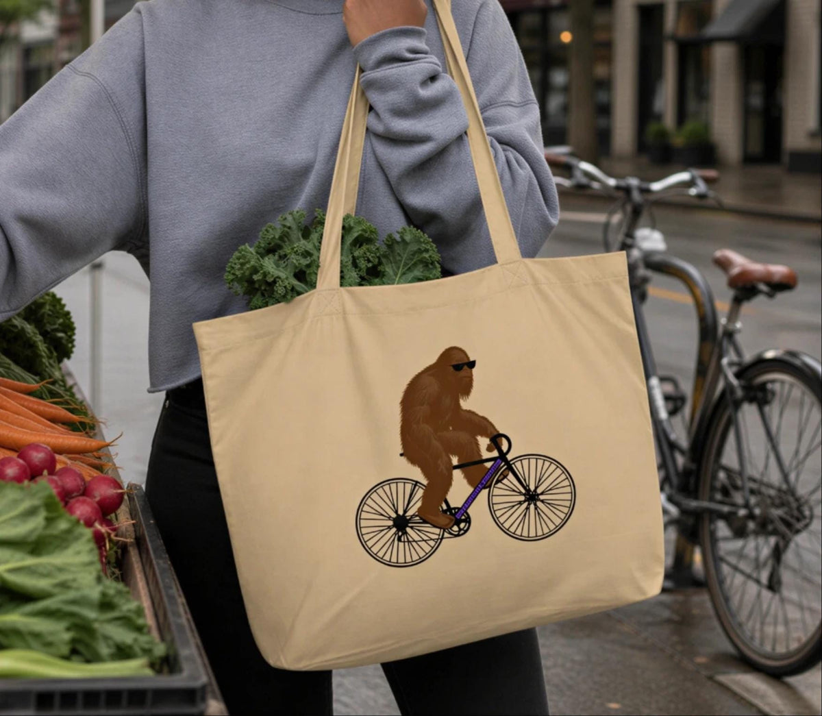 A person is holding a beige tote bag with a brown cartoon gorilla riding a bicycle printed on it.