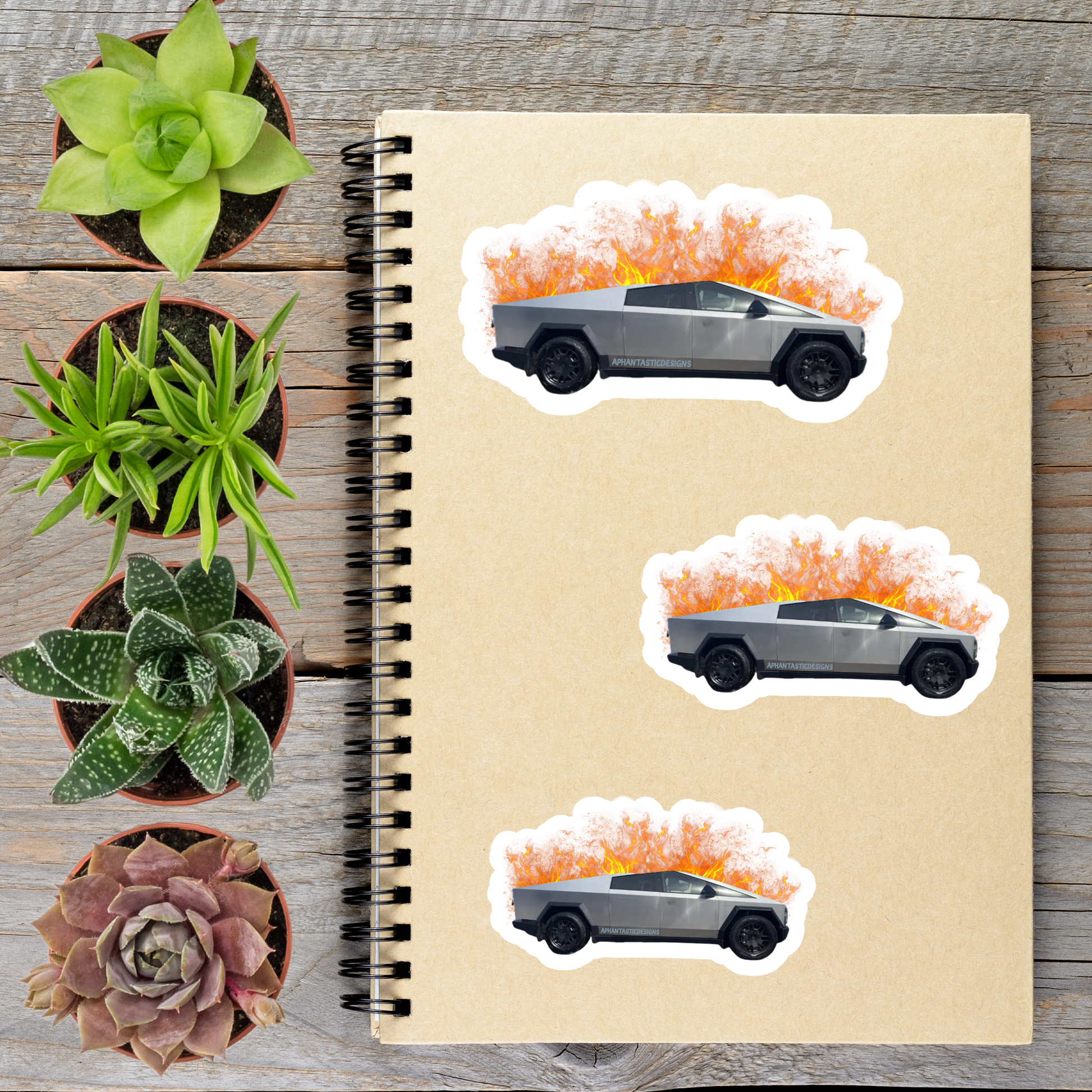 a spiral notebook with three stickers on it, depicting a silver truck with flames on top. The stickers are arranged in a row on the notebook, with the largest sticker on the left, the middle sticker slightly smaller, and the smallest sticker on the right. The notebook is placed on a wooden surface, with several potted plants surrounding it.