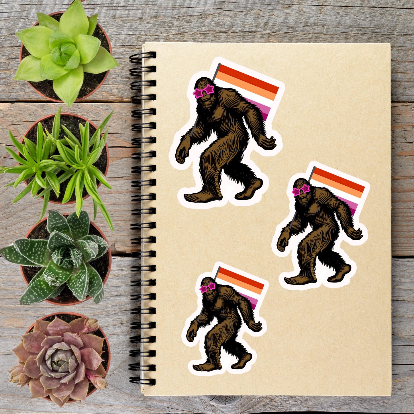 a notebook with stickers of three Sasquatch-like creatures, each wearing sunglasses and carrying a flag, alongside several potted plants on a wooden surface.