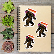 a notebook with stickers of three Sasquatch-like creatures, each wearing sunglasses and carrying a flag, alongside several potted plants on a wooden surface.
