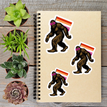 a notebook with stickers of three Sasquatch-like creatures, each wearing sunglasses and carrying a flag, alongside several potted plants on a wooden surface.