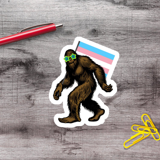a sticker of a cartoon Sasquatch wearing sunglasses and holding a flag, along with a red pen and yellow paper clips on a wooden surface.