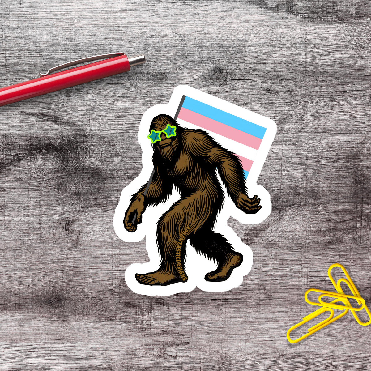 a sticker of a cartoon Sasquatch wearing sunglasses and holding a flag, along with a red pen and yellow paper clips on a wooden surface.