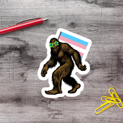 a sticker of a cartoon Sasquatch wearing sunglasses and holding a flag, along with a red pen and yellow paper clips on a wooden surface.
