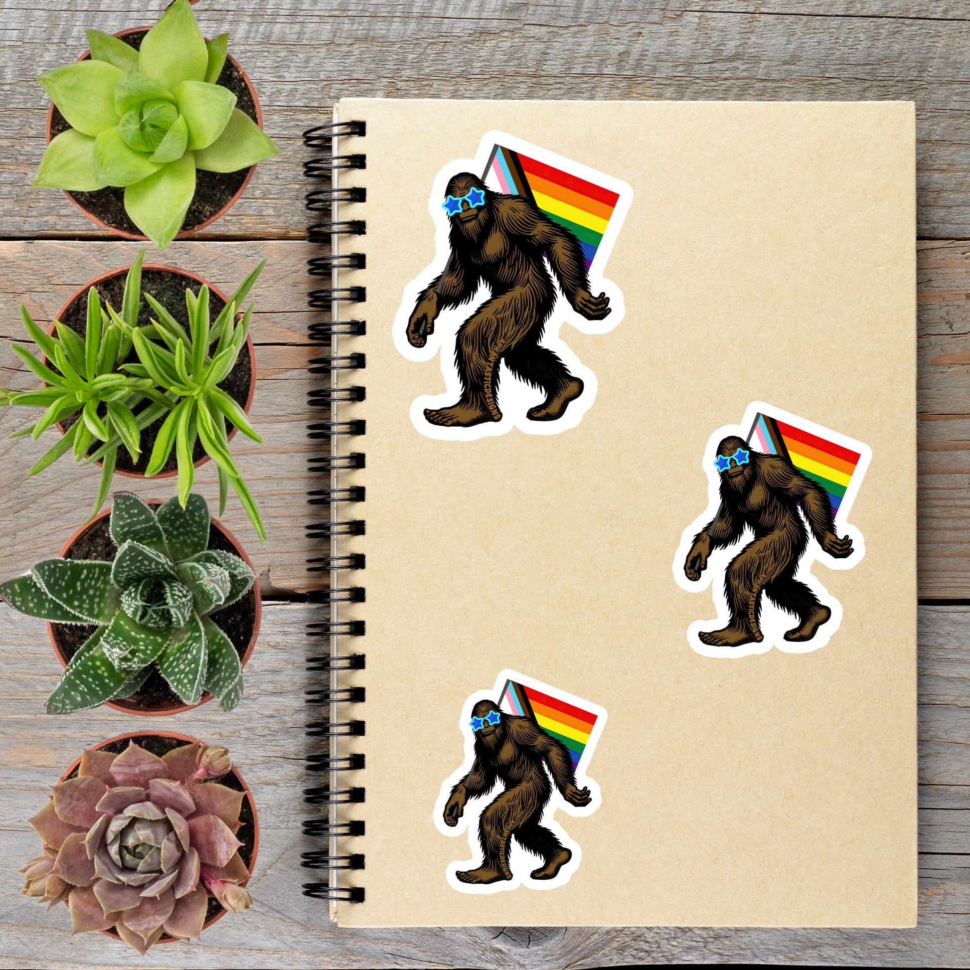 a spiral notebook with stickers of a Sasquatch wearing a rainbow flag, surrounded by potted plants and a wooden surface.