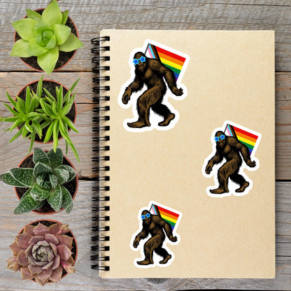 a spiral notebook with stickers of a Sasquatch wearing a rainbow flag, surrounded by potted plants and a wooden surface.