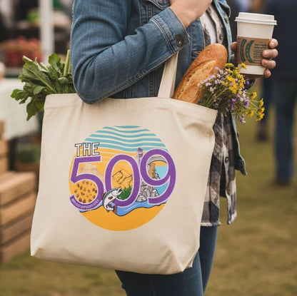 A woman is standing in a field, holding a white tote bag with a colorful graphic and text on it, and a cup of coffee in her hand.