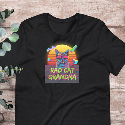 A black t-shirt with a graphic design featuring a cat wearing sunglasses and the text "RAD CAT GRANDMA" against a colorful sunset background.
