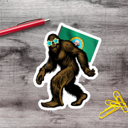 a sticker of a cartoon Sasquatch wearing sunglasses and holding a flag, along with a red pen and yellow paper clips on a wooden surface.