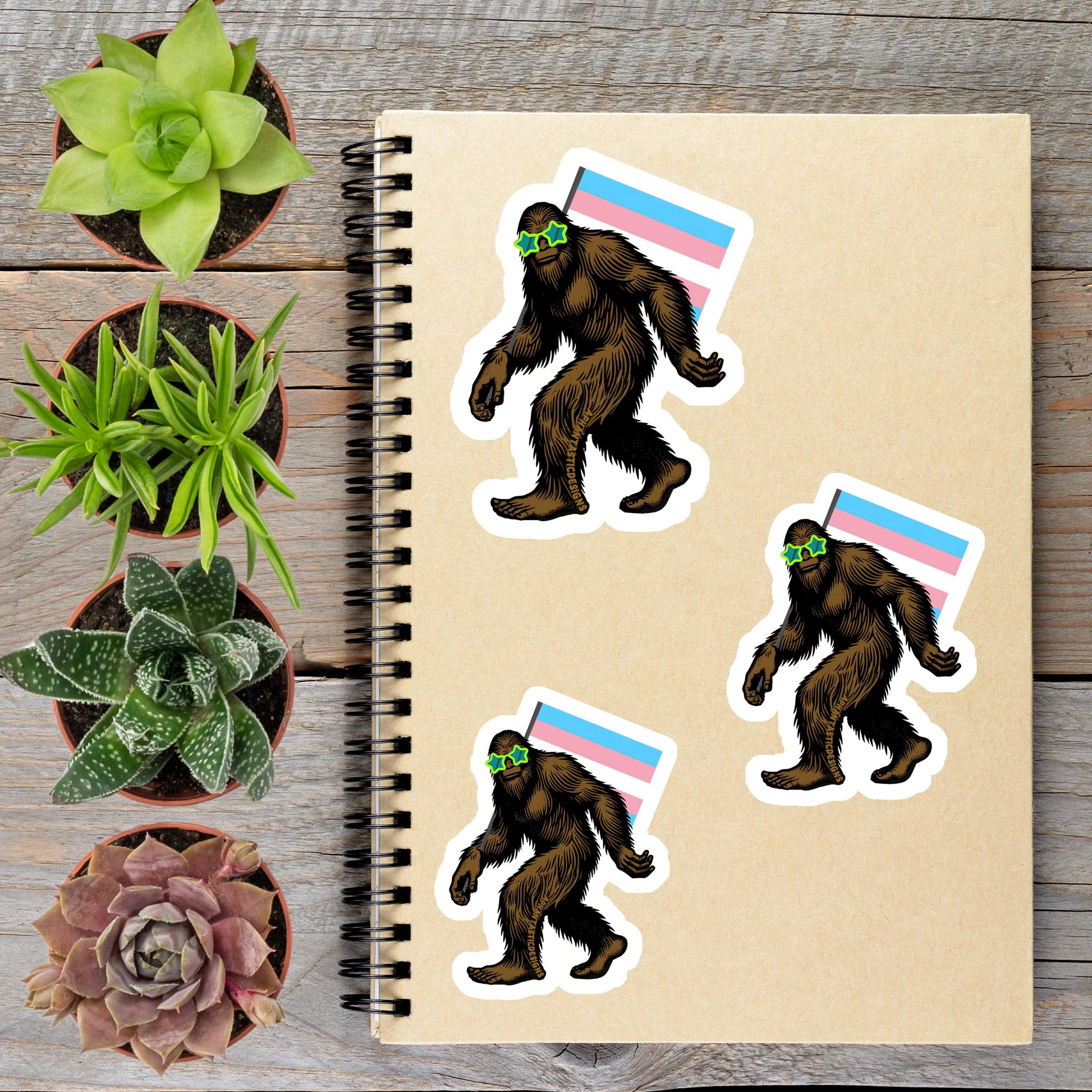 a spiral notebook with stickers of a Sasquatch wearing a pink, blue, and white flag, surrounded by potted plants and a wooden surface.