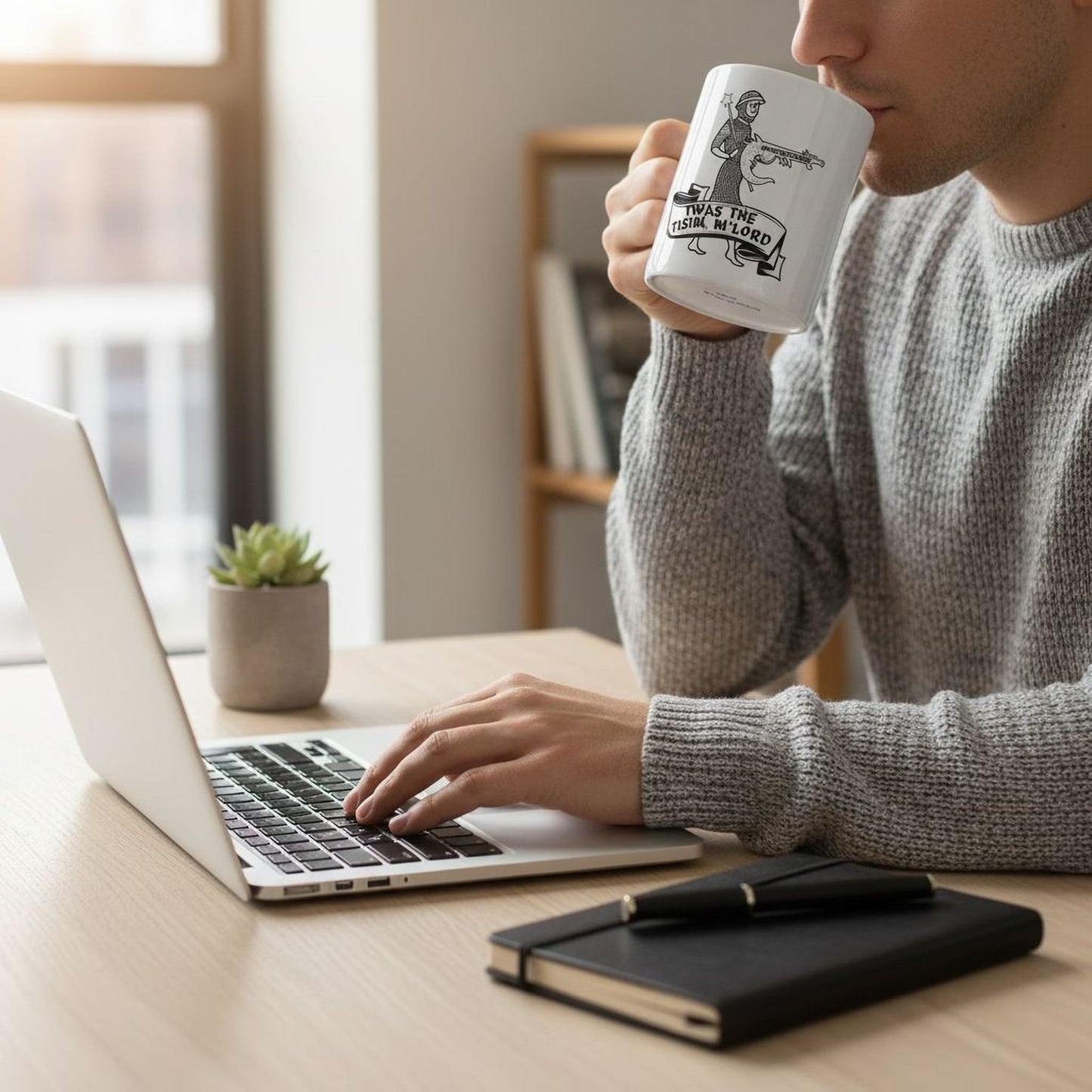 A person is sitting at a desk, working on a laptop while drinking from a mug. The mug has a graphic design on it.