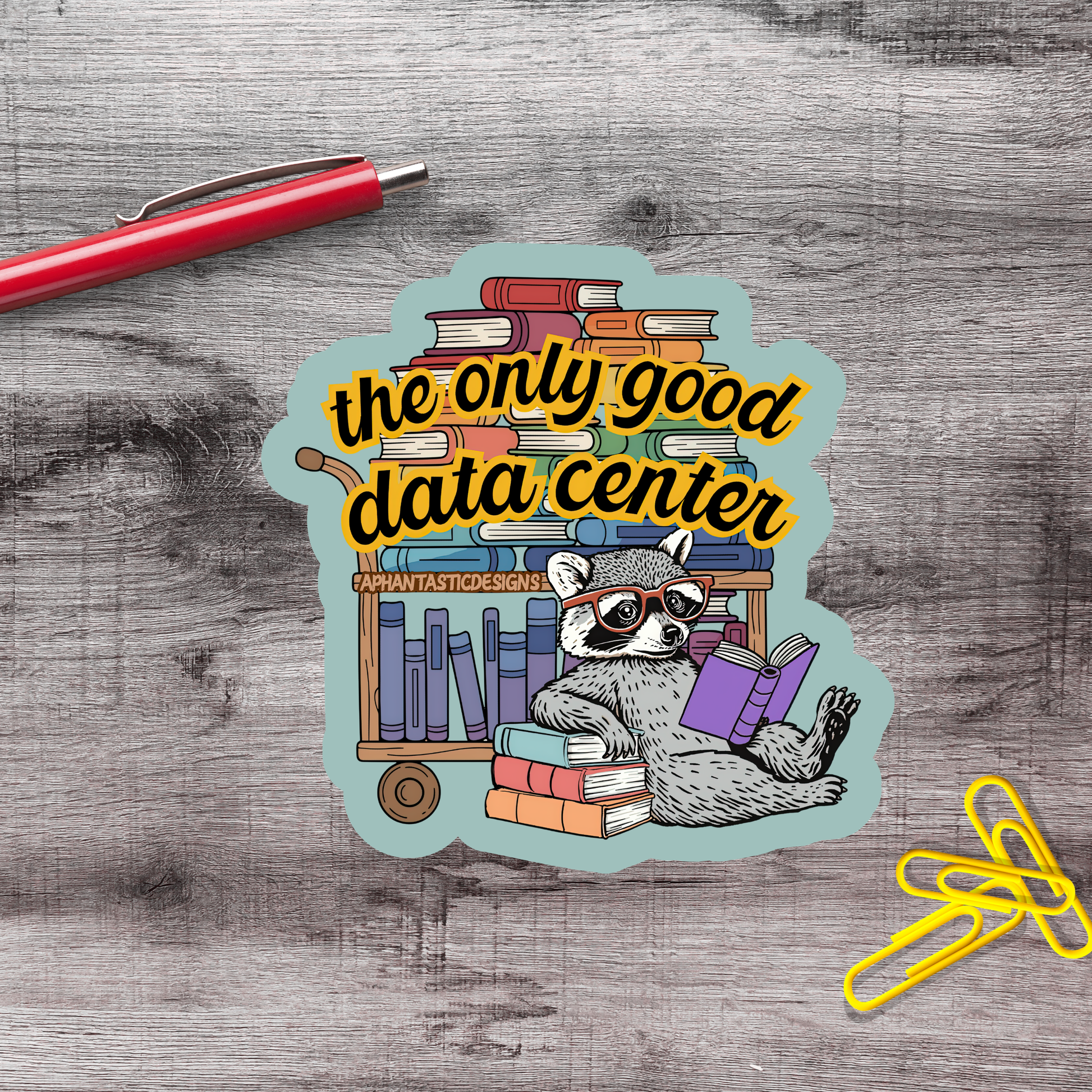 A sticker with a raccoon sitting on a bookshelf surrounded by books, with a red pen and yellow clips on a wooden surface.