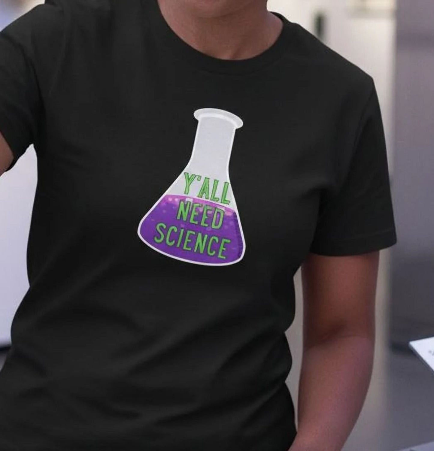 A woman wearing a black t-shirt with a graphic of a laboratory flask and the text "Y'ALL NEED SCIENCE" is holding a glass beaker filled with purple liquid.