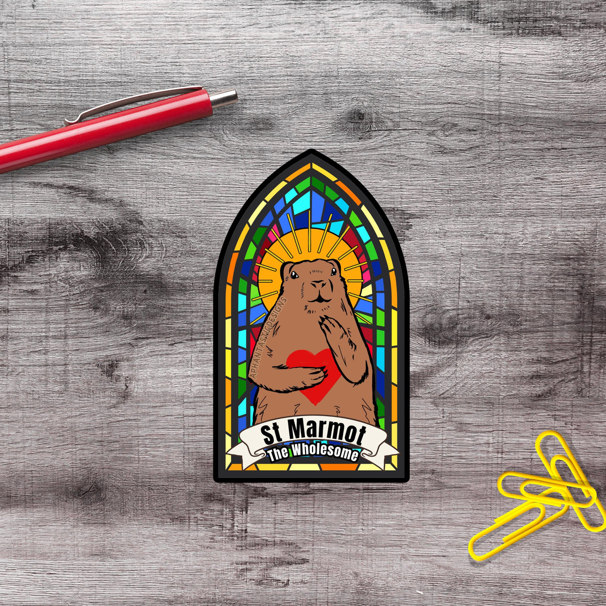 A sticker featuring a cartoon image of a brown bear with a heart, surrounded by a colorful stained glass window, is placed on a wooden surface next to a red pen and yellow paper clips.