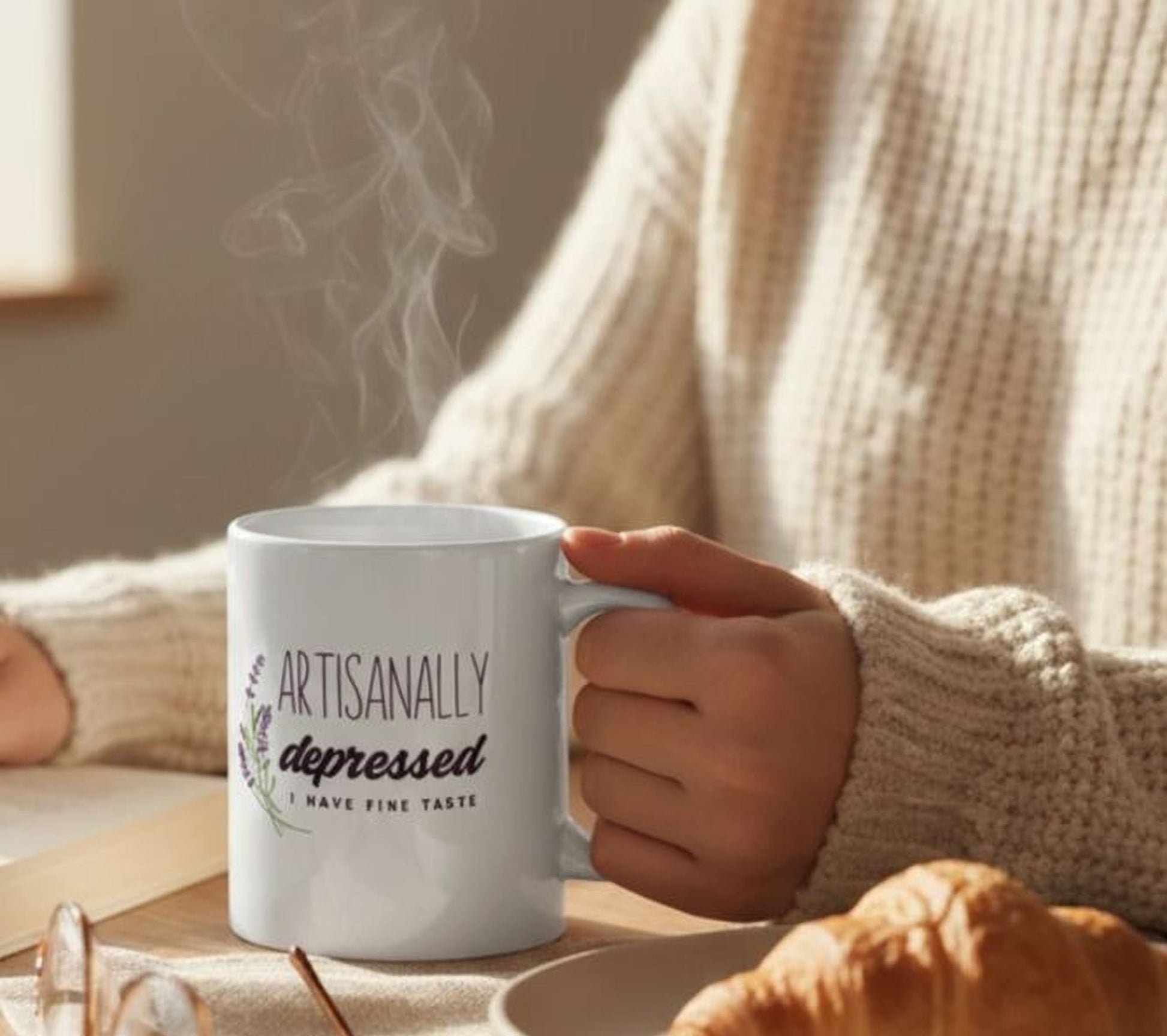 A person is holding a white mug with the text "Artistically Depressed I have fine taste" printed on it, while sitting at a table with a plate of food and a pastry.