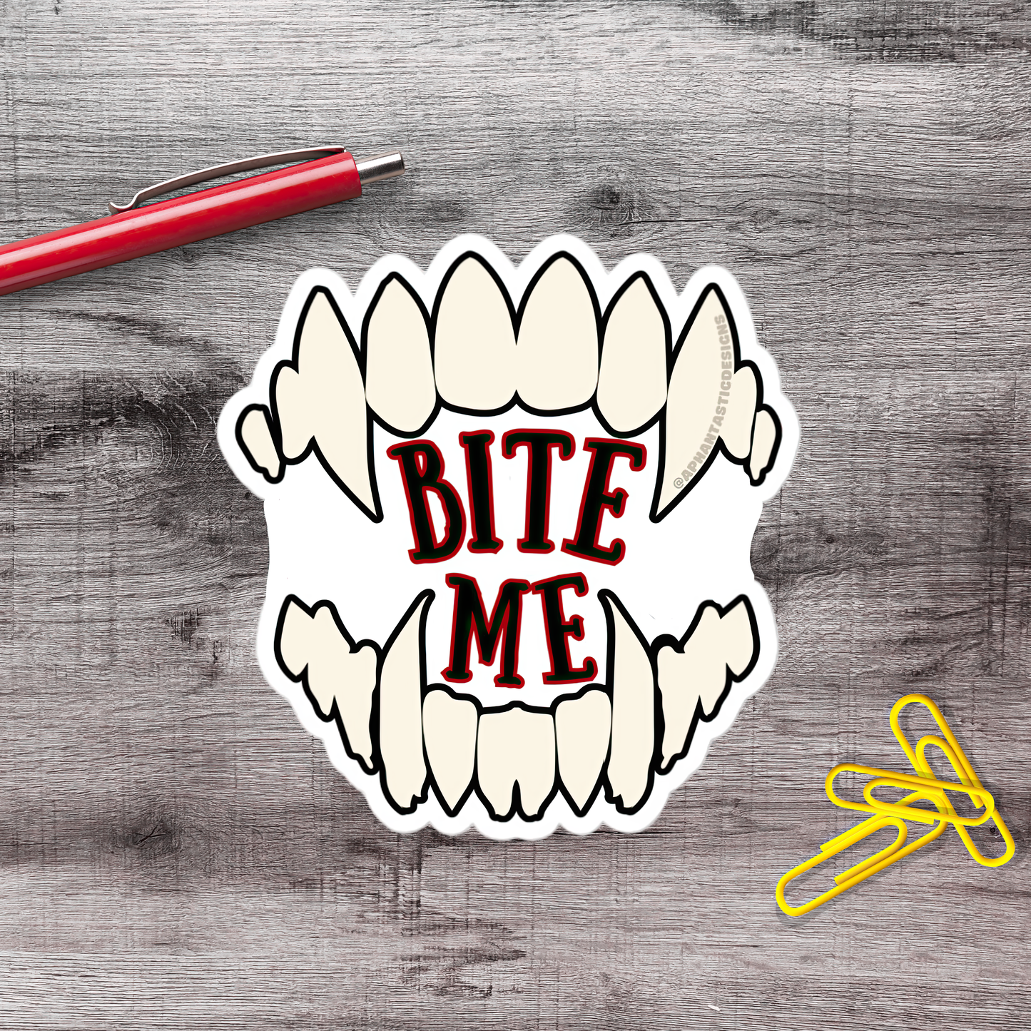 a sticker with the text "BITE ME" in bold red letters, which is placed on a wooden surface next to a red pen and a pile of yellow paper clips.
