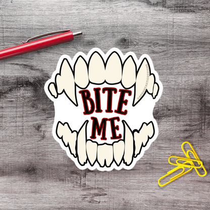 a sticker with the text "BITE ME" in bold red letters, which is placed on a wooden surface next to a red pen and a pile of yellow paper clips.