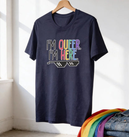 A navy blue t-shirt with the text "I'm queer. I'm here" printed on it, along with a graphic of a pair of sunglasses.