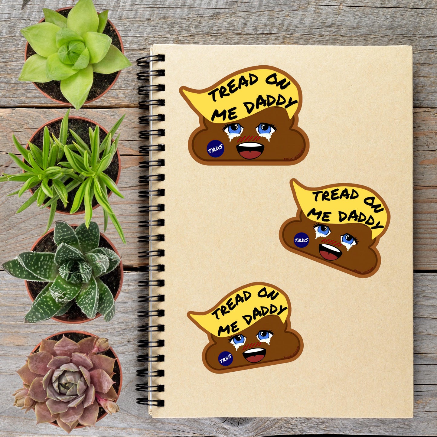 a spiral-bound notebook with three stickers on the cover, each depicting a cartoon character with a humorous caption. The stickers are arranged on the notebook, which is placed on a wooden surface alongside several potted plants.