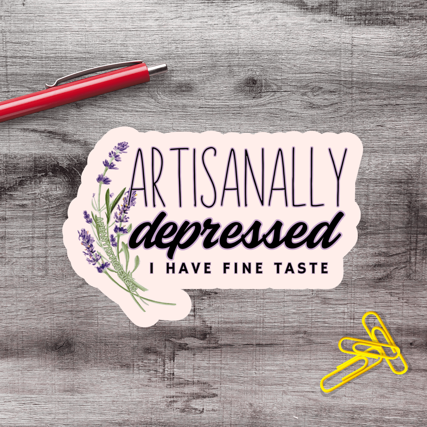 a sticker with the text "Artisanally Depressed I HAVE FINE TASTE" and a red pen on a wooden surface.