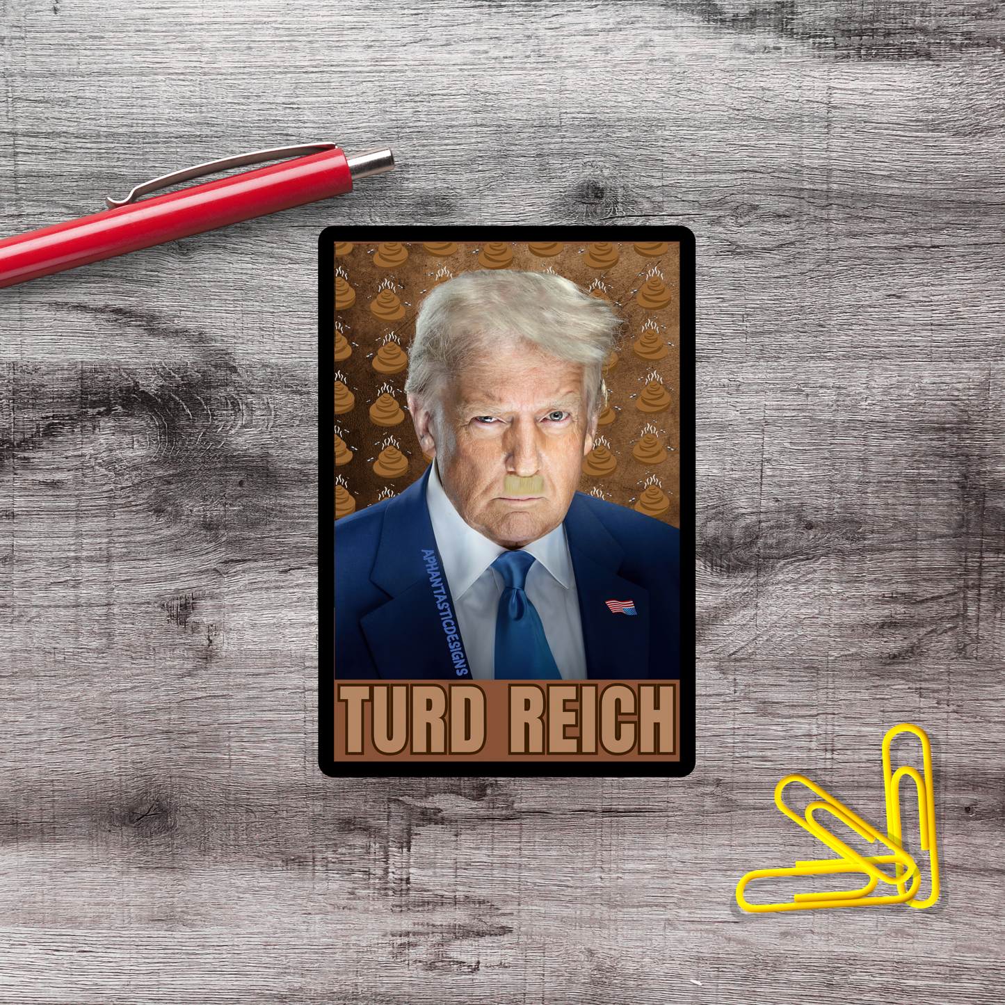 A political campaign card featuring a man with blonde hair and a blue suit, with the name 'TURD REICH' printed on it. The card is placed on a wooden surface, with a red pen and yellow paper clips nearby.
