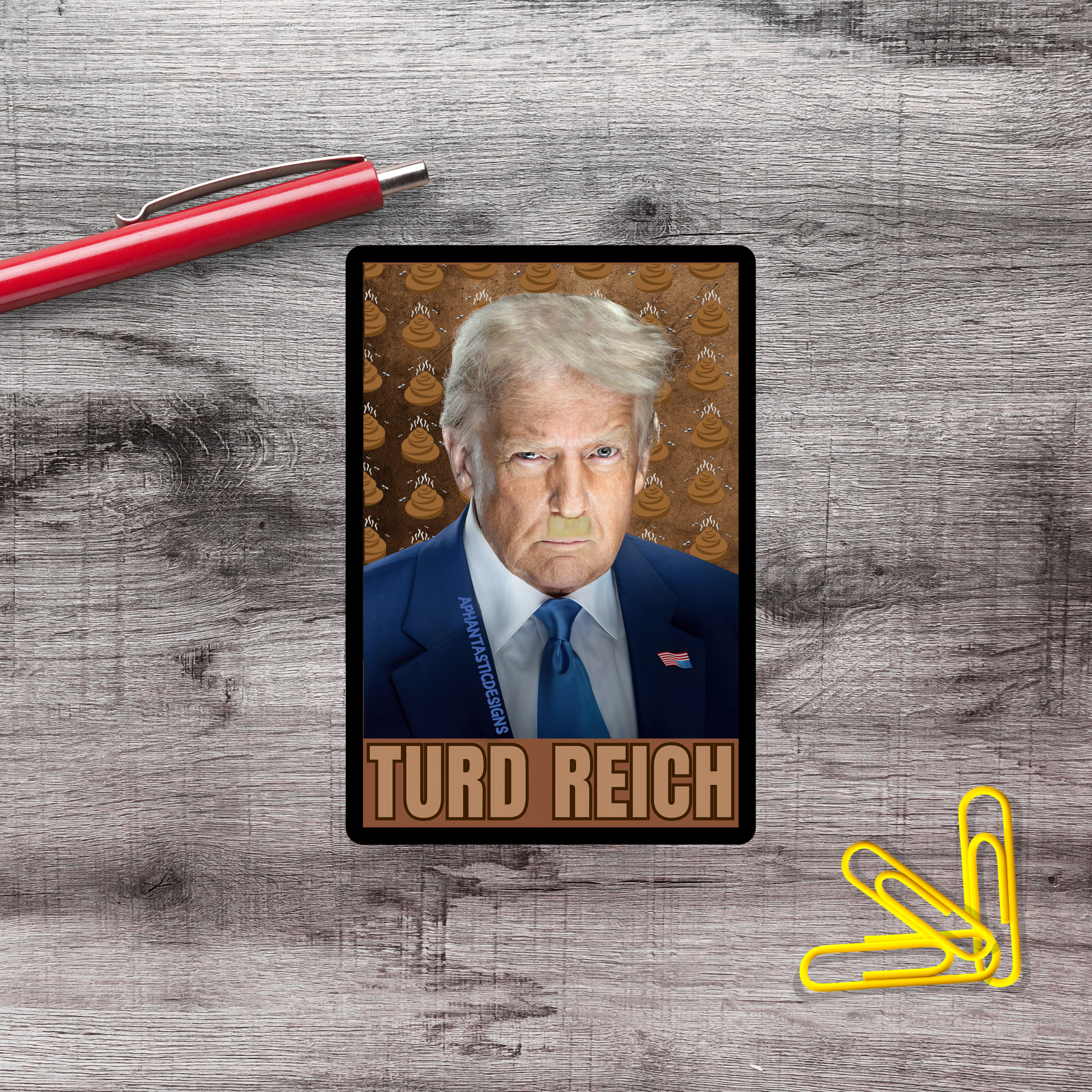 A political campaign card featuring a man with blonde hair and a blue suit, with the name 'TURD REICH' printed on it. The card is placed on a wooden surface, with a red pen and yellow paper clips nearby.