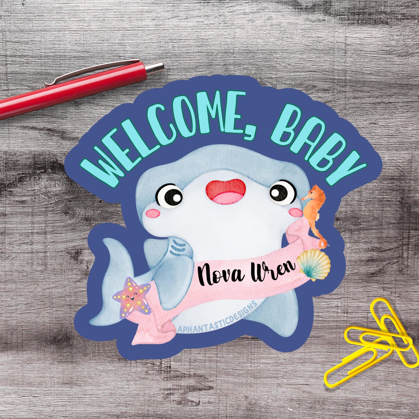 a welcome sign with a cartoon shark and a name tag, placed on a wooden surface alongside a red pen and yellow paper clips.