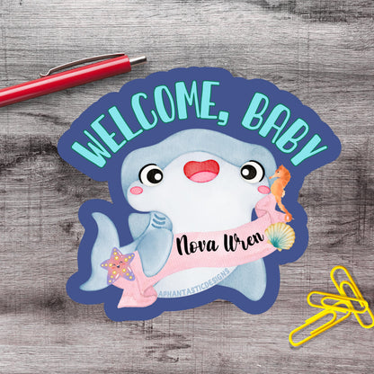 a welcome sign with a cartoon shark and a name tag, placed on a wooden surface alongside a red pen and yellow paper clips.
