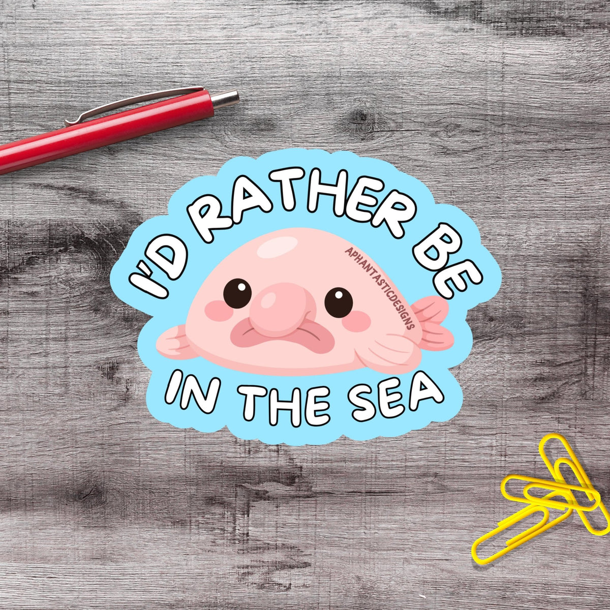 a sticker with a pink fish and the text "I'd rather be in the sea" on a wooden surface.