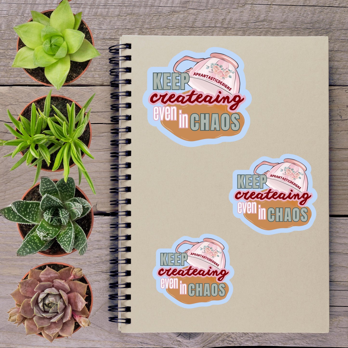 a spiral notebook with three stickers on it, along with several potted plants arranged on a wooden surface.