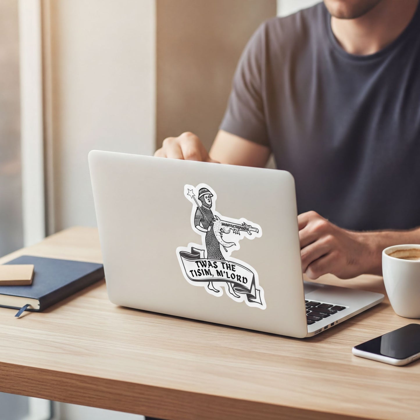A person is sitting at a desk using a laptop with a sticker on it, which features a cartoon character holding a sign that says "THAT IS THE TISIMM M'LORD".