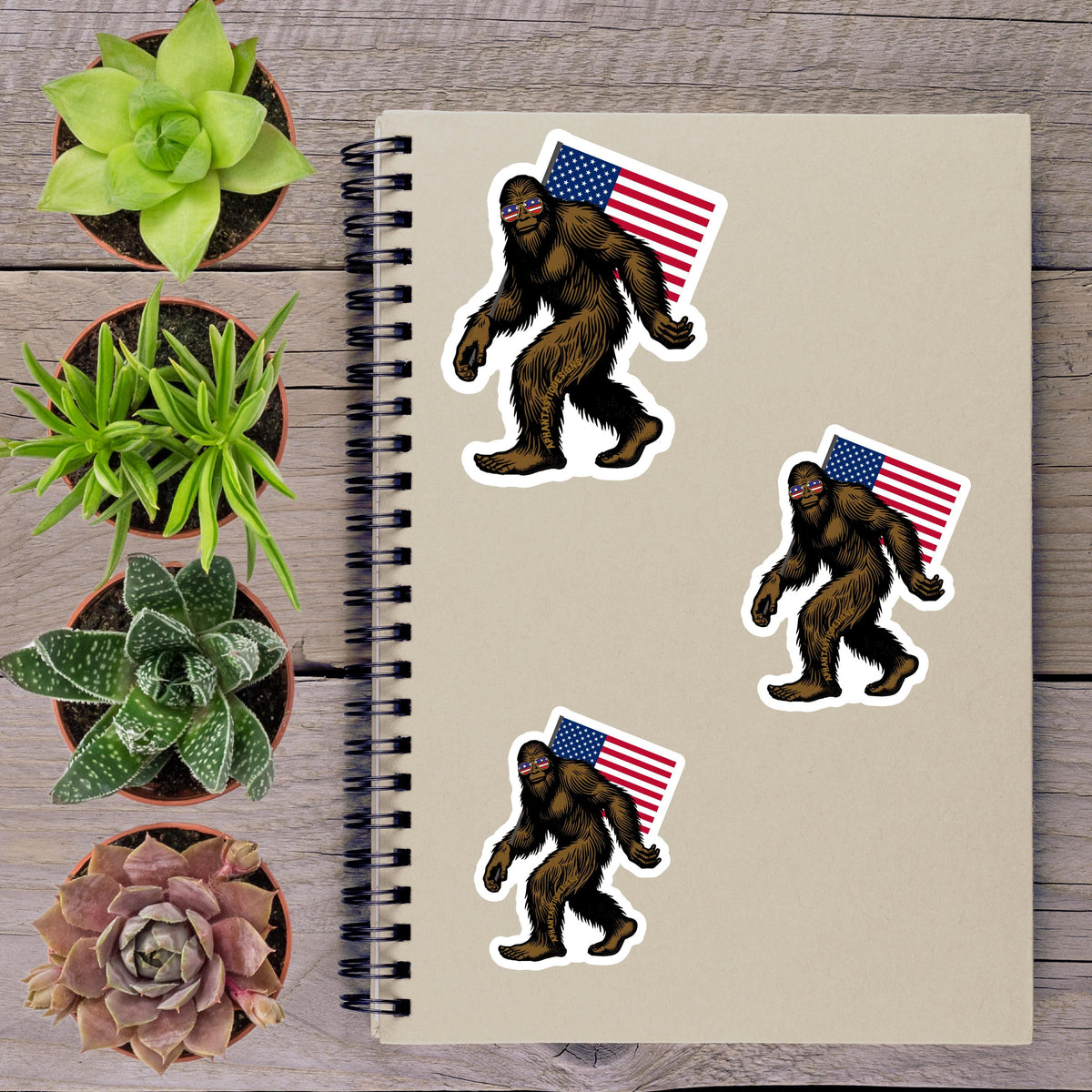 a spiral notebook with stickers of Sasquatch figures and an American flag, along with several potted plants arranged on a wooden surface.