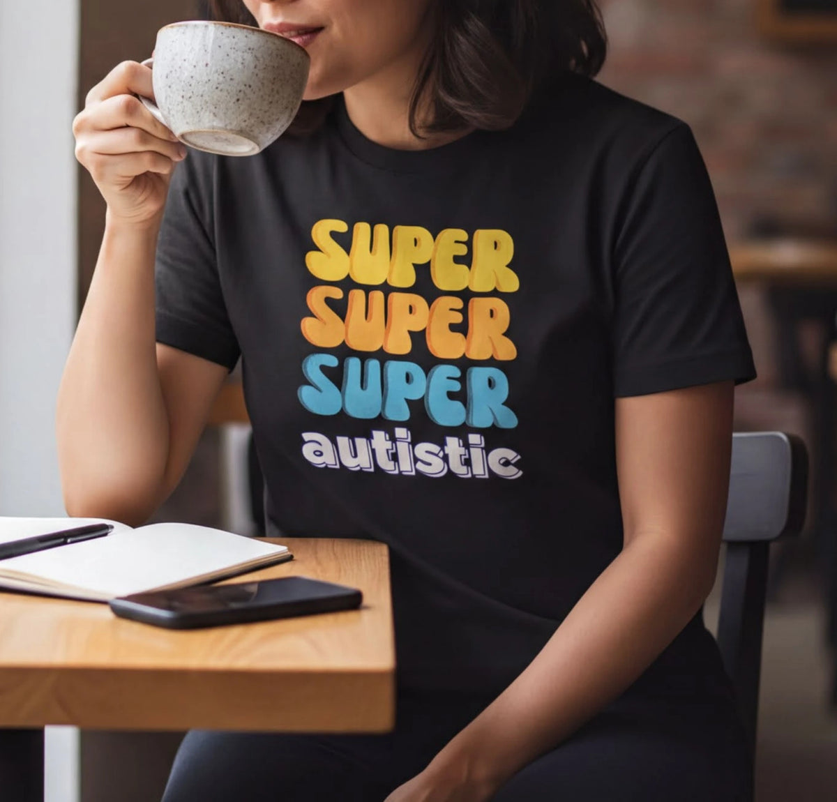 A woman wearing a black t-shirt with text on it is sitting at a table, sipping from a cup and looking out a window.