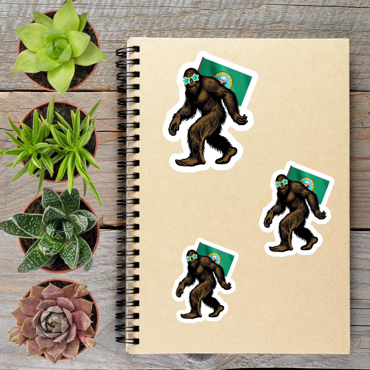 a spiral-bound notebook with stickers of cartoon-like Sasquatch figures, surrounded by potted plants and a wooden surface.