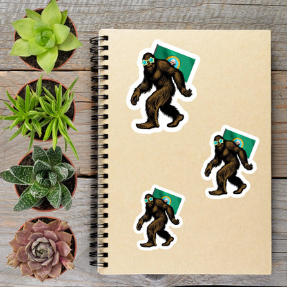 a spiral-bound notebook with stickers of cartoon-like Sasquatch figures, surrounded by potted plants and a wooden surface.