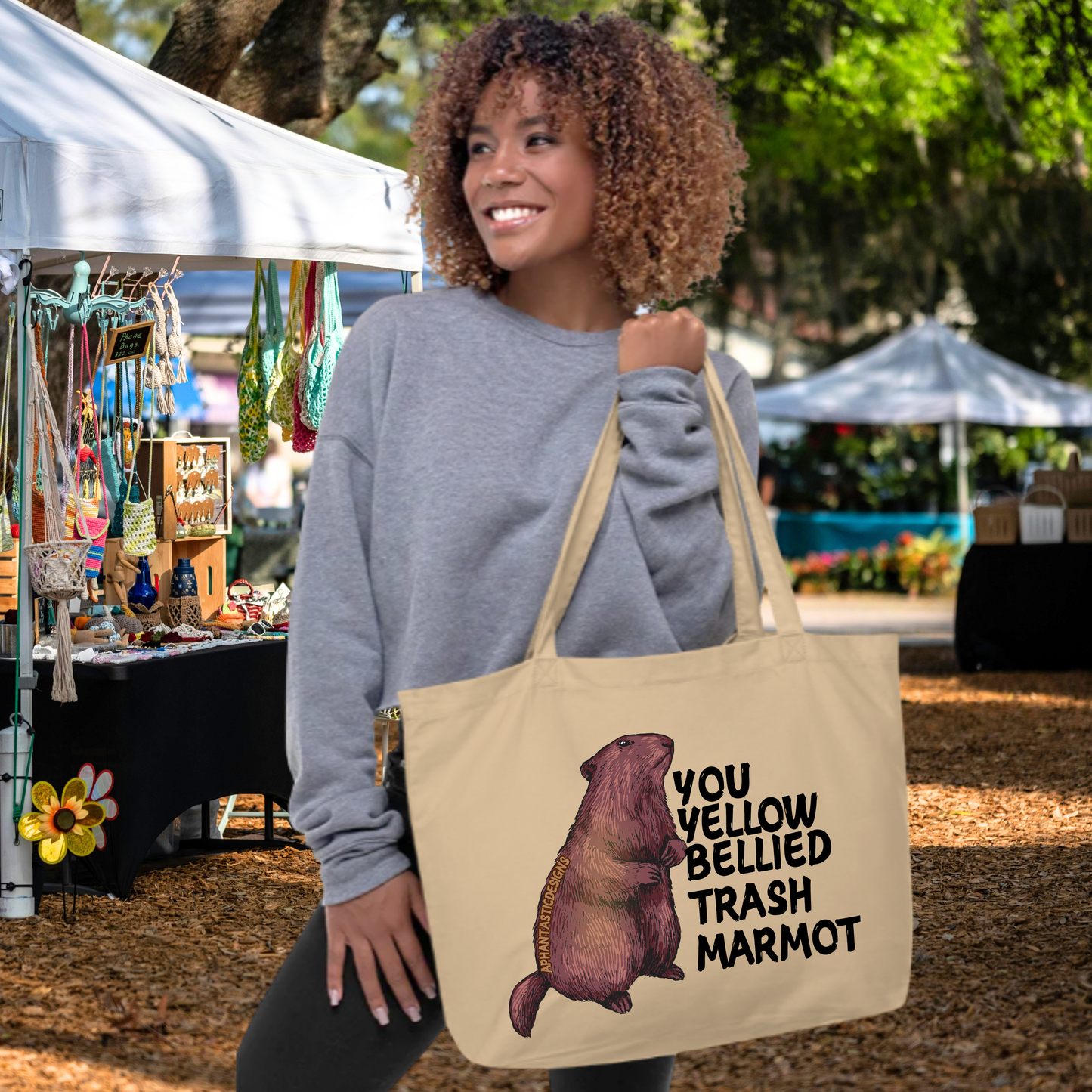A woman with curly hair is standing in front of a tent, holding a large tote bag with a printed design of a marmot.